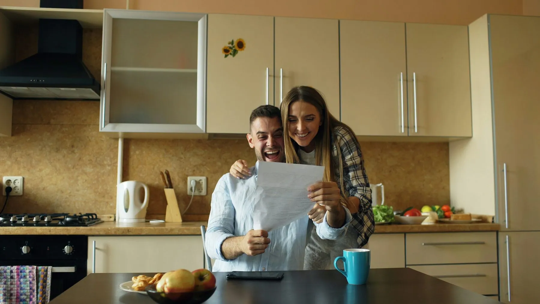 Attractive happy couple recieve good news unfolding letter in the kitchen while have breakfast at home early morning