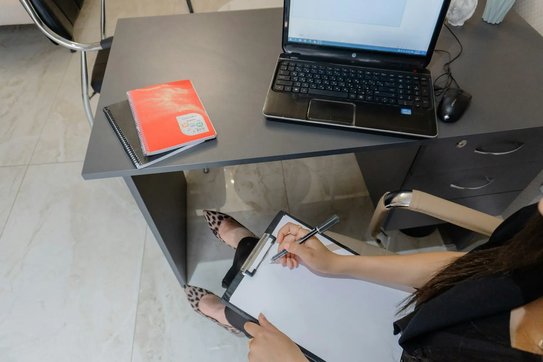 Person writing on clipboard at desk with laptop