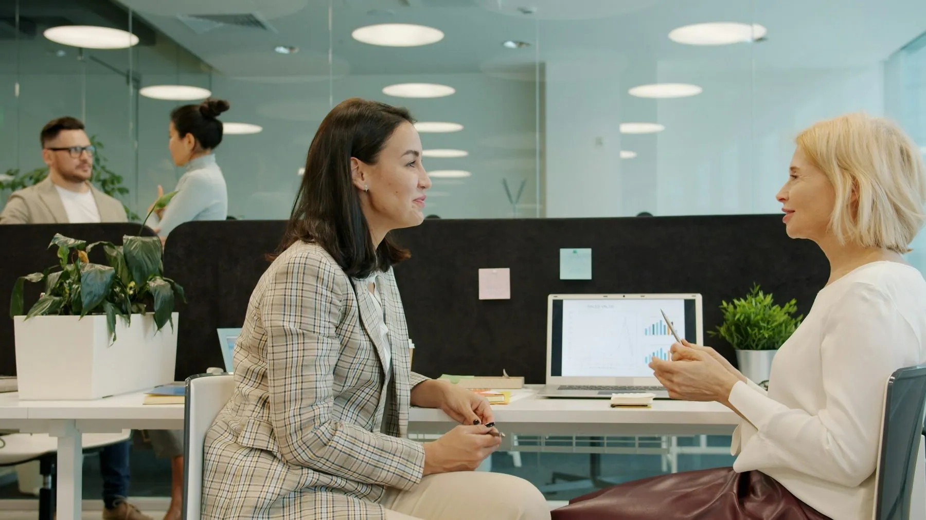 Cheerful women coworkers talking discussing work looking at laptop screen sitting at desk in open space office room. Peo